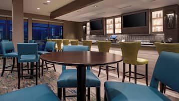 A hotel lobby with a large round table, blue and yellow chairs, a TV, and a bar area with bottles.