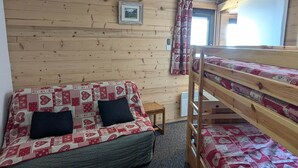 A room with a bunk bed, a single bed with patterned bedding, a small wooden stool, a framed picture on the wall, and a window with curtains.