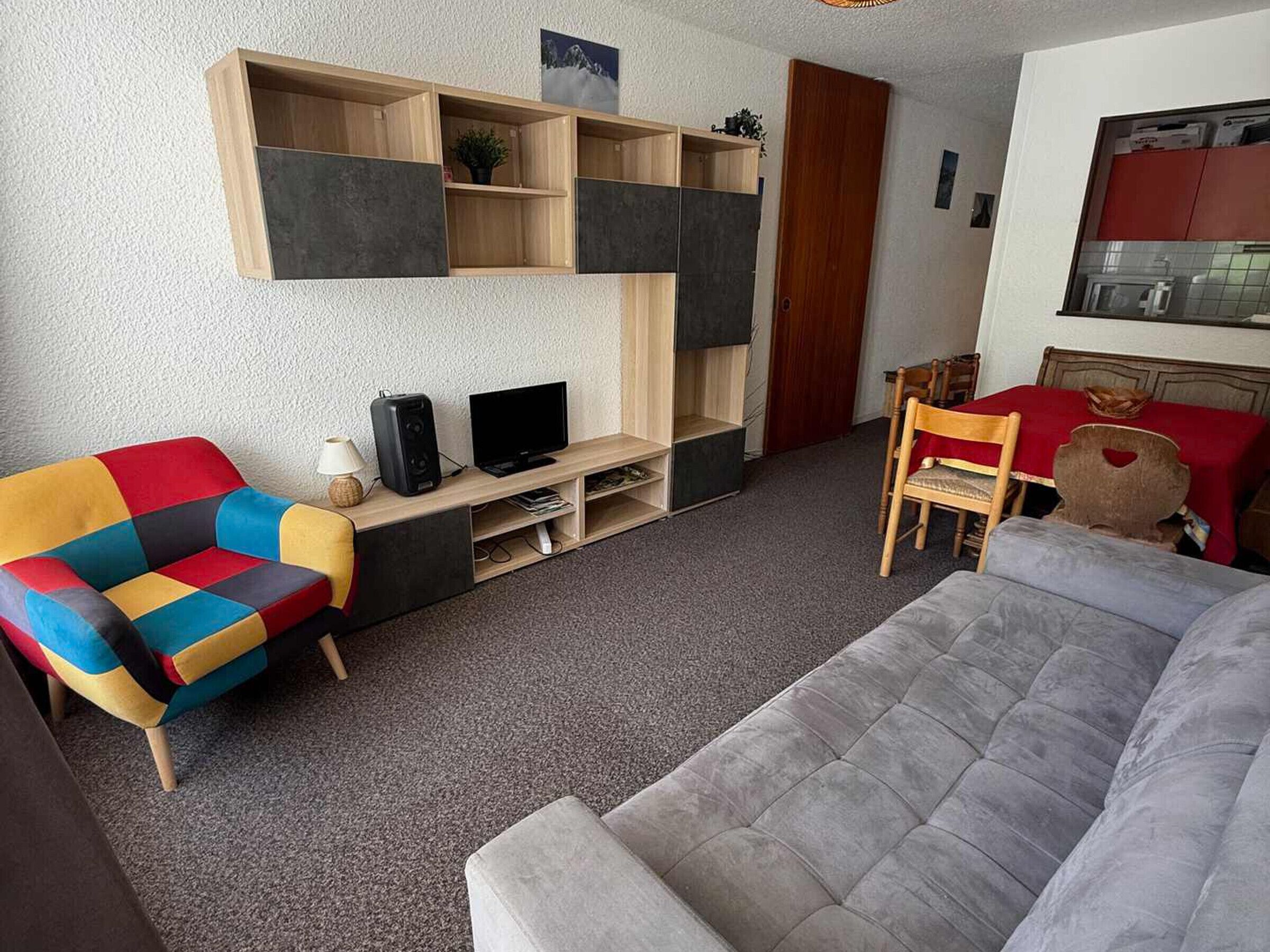 Ein Wohnzimmer mit einer Couch, einem bunten Sessel, einem TV-Schrank und einem Essbereich mit Tisch und Stühlen.