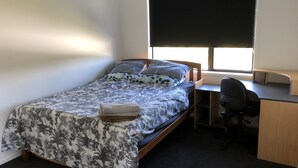 A single bed with a floral bedspread, a bedside table, a desk with a chair, and a window with blinds.