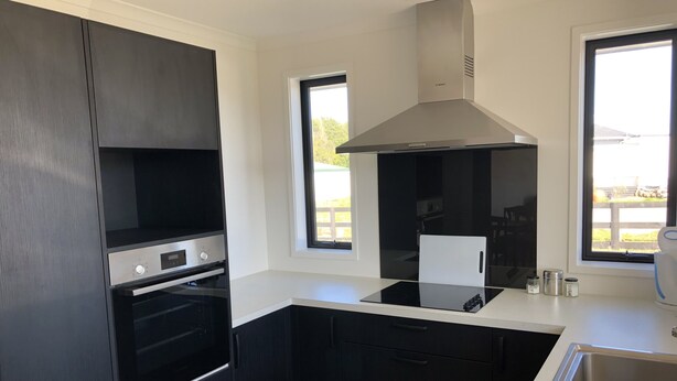 A modern kitchen with black cabinets, a stainless steel range hood, a built-in oven, a white countertop, and a window providing a view of the outdoors.