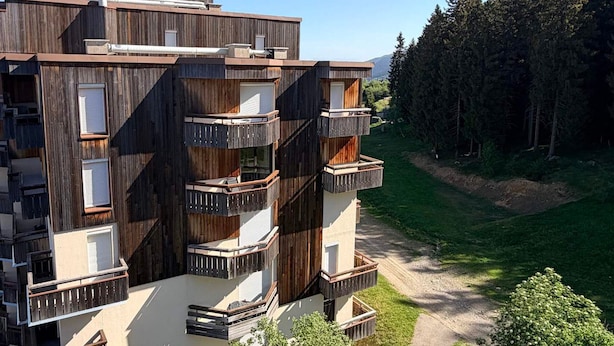 A multi-story building with balconies, a flat roof, and a surrounding forest.