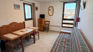 A room with a wooden bed, a sofa, a table with chairs, a TV on a wooden cabinet, and a window with a view of buildings.