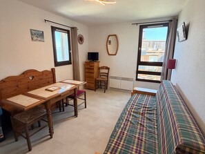 Ein Zimmer mit einem hölzernen Bett, einem Sofa, einem Tisch mit Stühlen, einem Fernseher auf einer Holzkommode und einem Fenster mit Blick auf Gebäude.