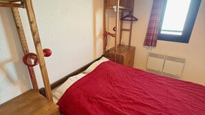 A bedroom with a wooden bunk bed, a red bedspread, a wardrobe, a mirror, and a window with curtains.