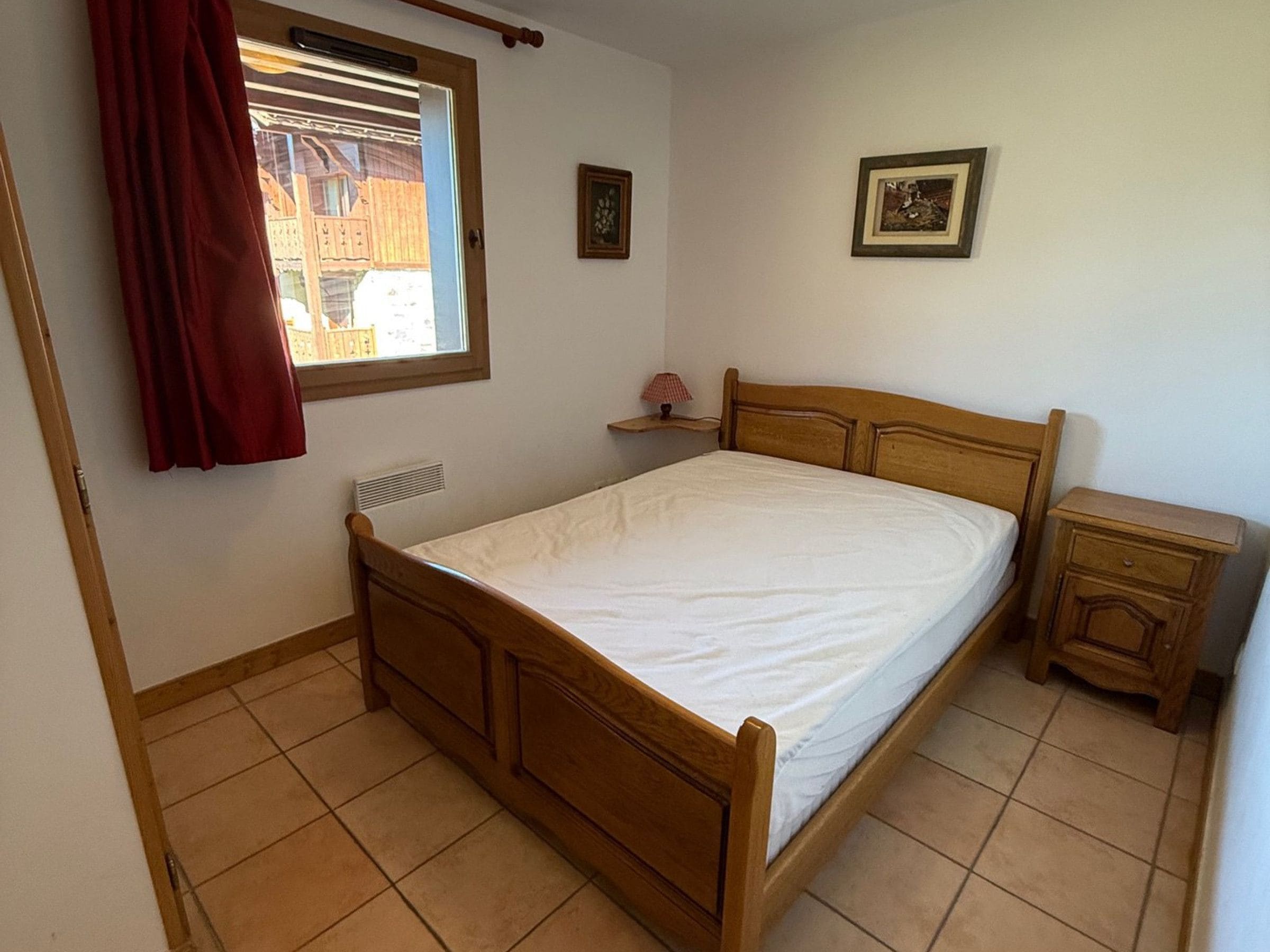 Ein Schlafzimmer mit einem hölzernen Bett, einem Nachttisch, einem Fenster mit rotem Vorhang und einem an der Wand hängenden Bild.