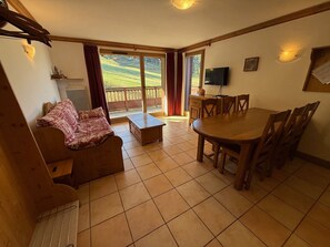 A living room with a sofa, a coffee table, a dining table with chairs, a TV, and a balcony with curtains. - 3-room luxury apartment at the foot of the slopes with private parking (Les Adrets-Prapoutel)