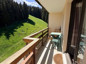 A balcony with a wooden railing, a table, and chairs, overlooking a green landscape with trees and a hill.