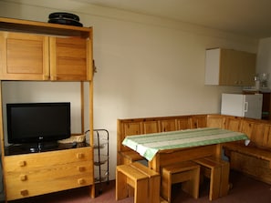 Quarto compacto com uma cama de madeira beliche, televisão, pequena cozinha e um banco.