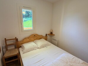 A single bed with white bedding, a wooden headboard, a small wooden chair, a bedside table with a lamp, and a window with a view of greenery.