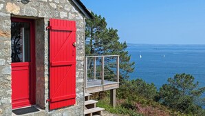Un bâtiment en pierre avec une porte rouge et une terrasse en bois donnant sur une étendue d’eau.