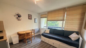 A compact living space with a wooden dining table, a sofa with storage drawers, a window with blinds, and a wall-mounted bulletin board.
