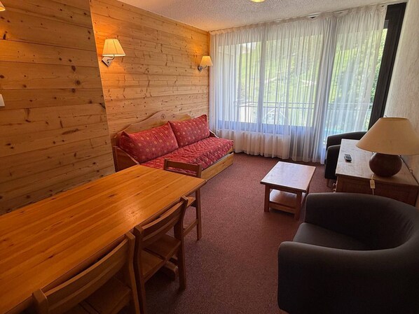 A room with a wooden wall, a sofa, a table, a chair, and a window with curtains.