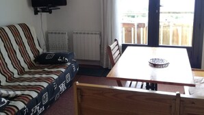 Een kamer met een gestreepte bank, een aan de muur bevestigde televisie, een eettafel met stoelen en uitzicht op een houten terras door een raam.