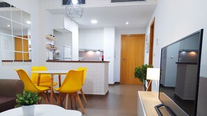 Un salon moderne avec une table ronde blanche, des chaises jaunes, un canapé marron, un téléviseur posé sur un meuble et un coin cuisine avec des placards blancs et une porte en bois.