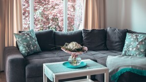 A modern living room with a grey sofa, floral cushions, a small white side table, and a vase of flowers.
