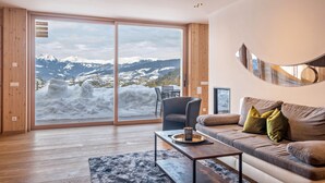 Una sala de estar moderna con un amplio ventanal corredero que ofrece vistas a un paisaje montañoso nevado, un sofá con cojines, una mesa de centro y un sillón.