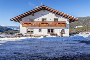 A two-story building with a balcony, a sign reading 'Tschagg - Hof,' and a snowy landscape in the background. - Beautiful Apartment “Aura Chalets Nr 1” with Mountain View, Wi-Fi & Garden (Kastelruth)