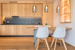 Une cuisine moderne avec des meubles en bois, des luminaires suspendus, un coin repas avec deux chaises et une étagère intégrée où sont rangés de la vaisselle.