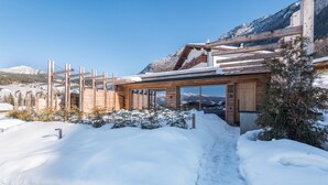A modern wooden building with large windows, surrounded by snow-covered ground and mountains in the background. - Beautiful Holiday Apartment "Aura Chalets No 4" with Mountain View, Wi-Fi & Garden (Kastelruth)