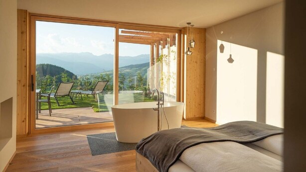 A bedroom with a large window, a bathtub, and a view of a green landscape. - Beautiful Holiday Apartment "Aura Chalets No 4" with Mountain View, Wi-Fi & Garden (Kastelruth)