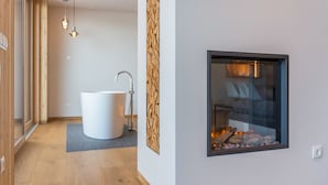 A modern bathroom with a white bathtub, a wooden wall panel, and a glass fireplace. - Beautiful Holiday Apartment "Aura Chalets No 4" with Mountain View, Wi-Fi & Garden (Kastelruth)