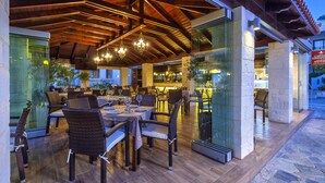 An outdoor dining area with wicker chairs and tables, wooden beams, and hanging lights. - Hotel Castle Suites (Platanias)