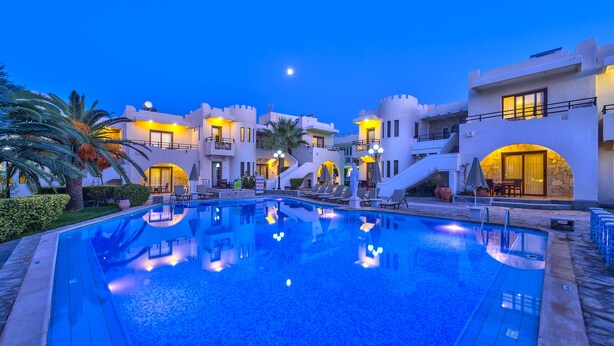 A pool area with a reflection of a building, palm trees, and a clear sky. - Hotel Castle Suites (Platanias)