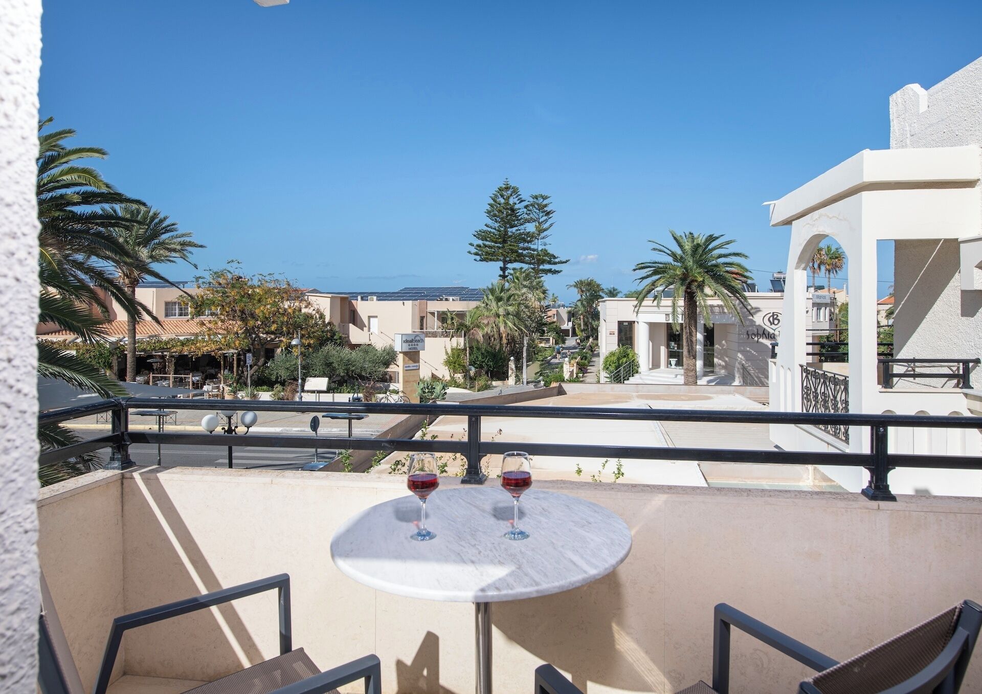 A balcony with two round tables, each with a wine glass, overlooking a street with buildings and palm trees.