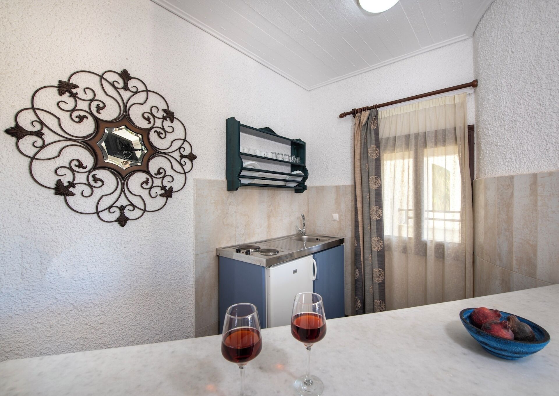 Uma cozinha pequena com pia, um detalhe decorativo na parede e janela com cortinas. Há dois copos de vinho tinto na bancada.