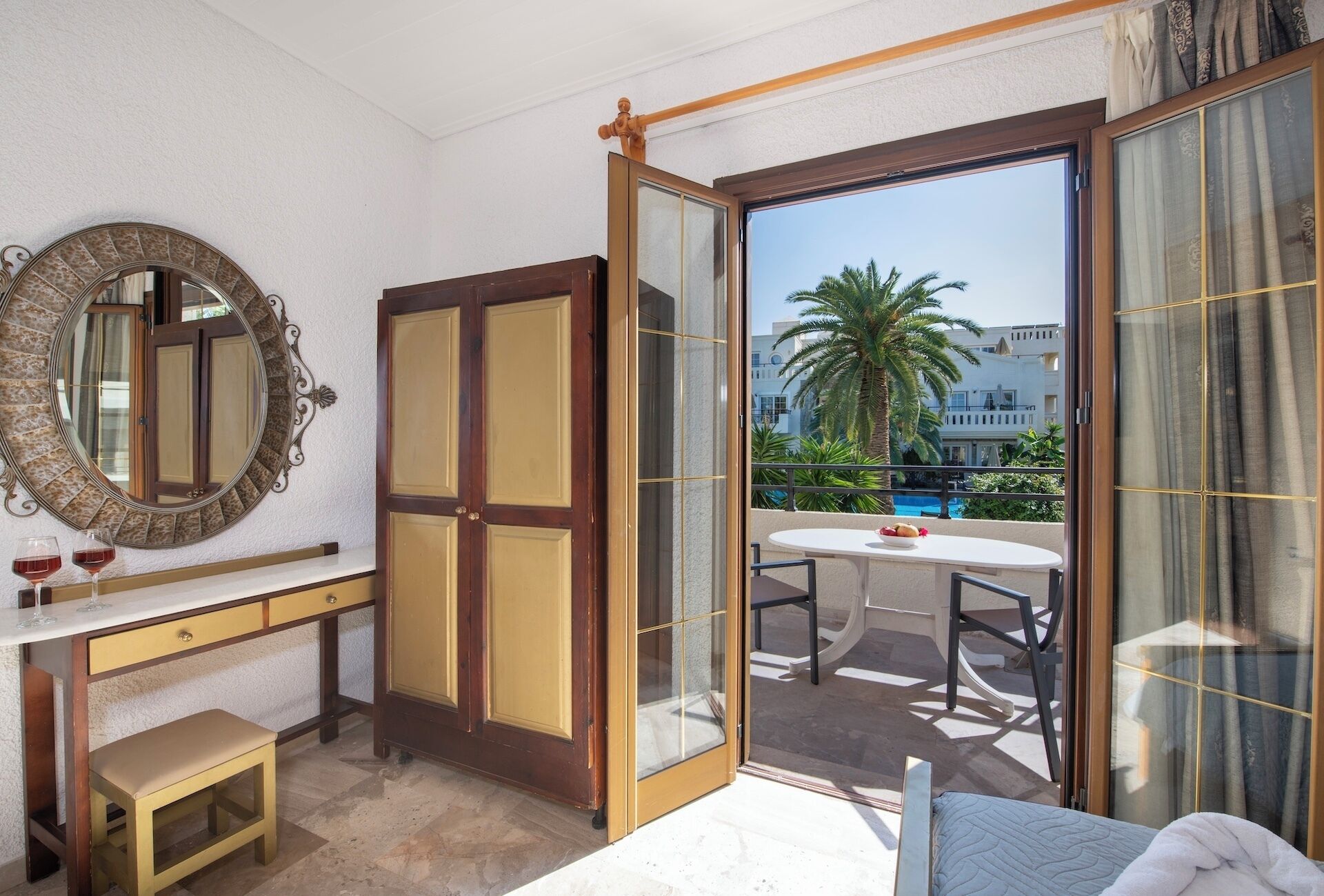 A hotel room with a balcony, a round mirror, a wooden wardrobe, a small table with chairs, and a bed.