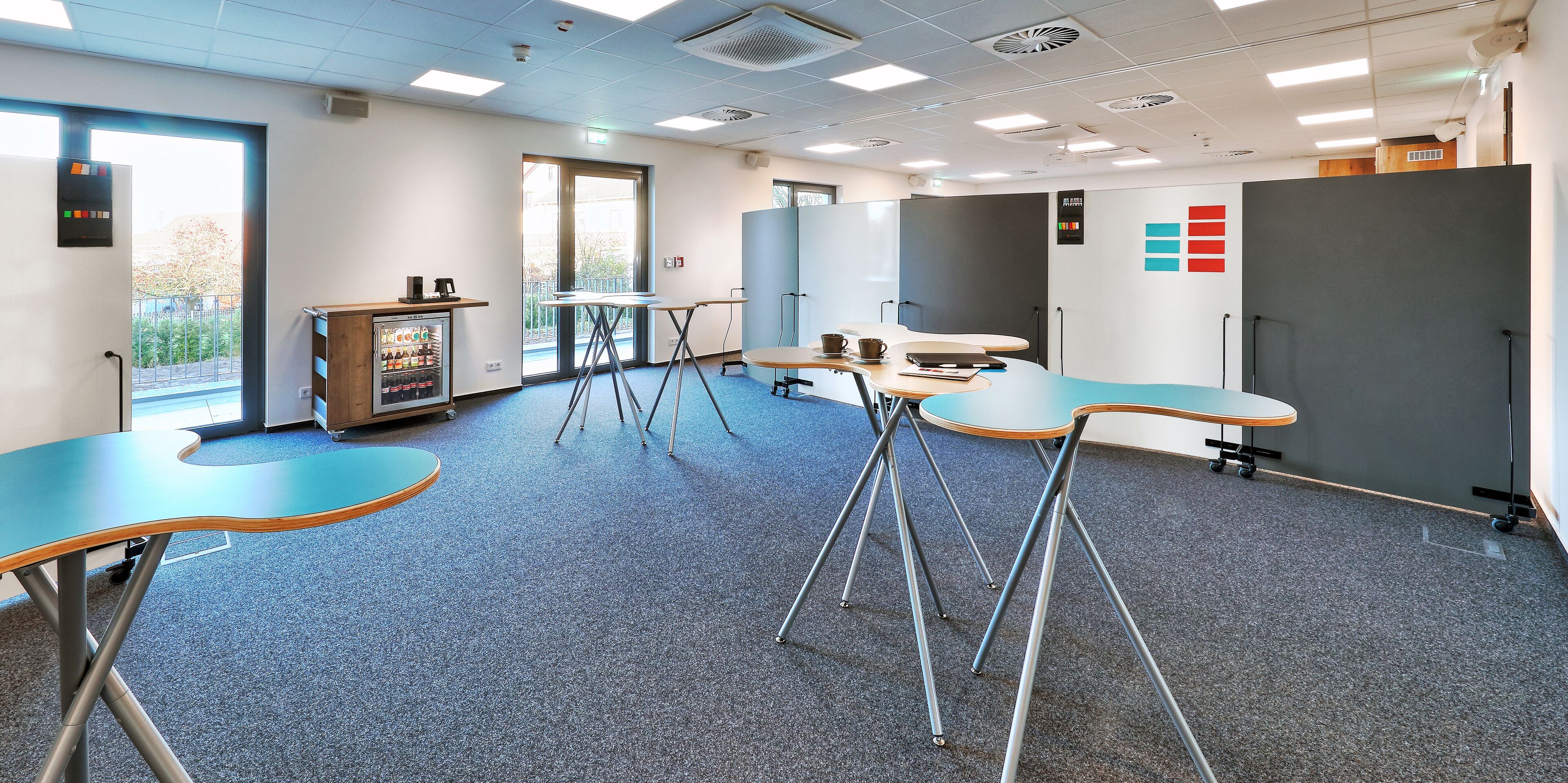 A modern office space with a blue carpet, several tables and chairs, a vending machine, and a large grey partition.