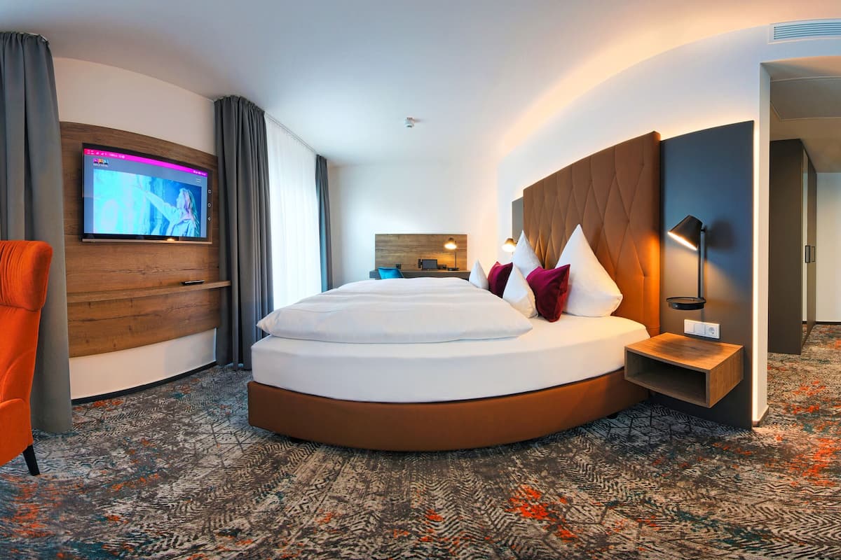 A modern hotel room with a curved bed, a sofa, an armchair, a TV, and a minibar.