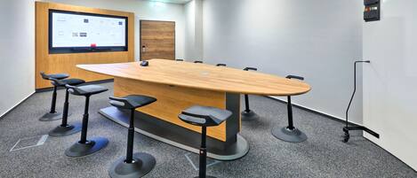 Un salón de conferencias con una mesa larga, cuatro taburetes altos, una pantalla de proyector y una pizarra.