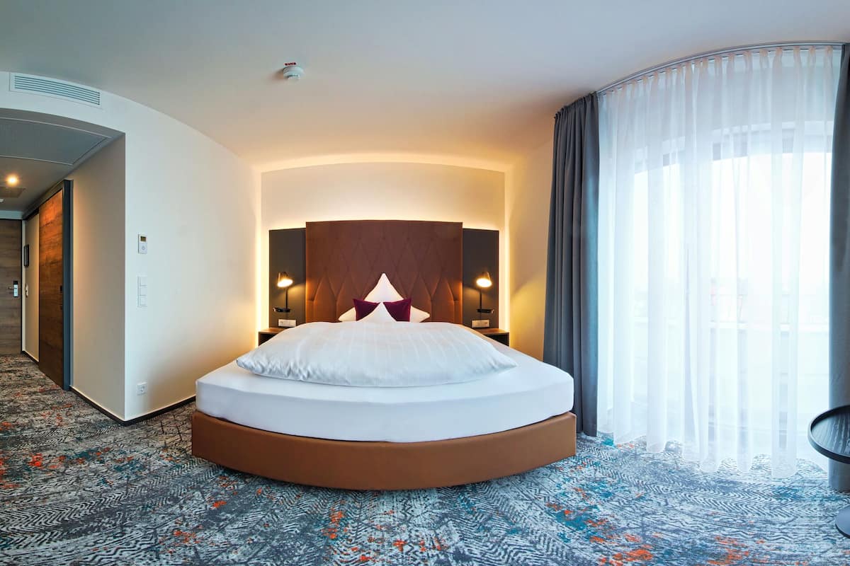 A modern hotel room with a curved bed, a TV mounted on the wall, and a seating area with two chairs.