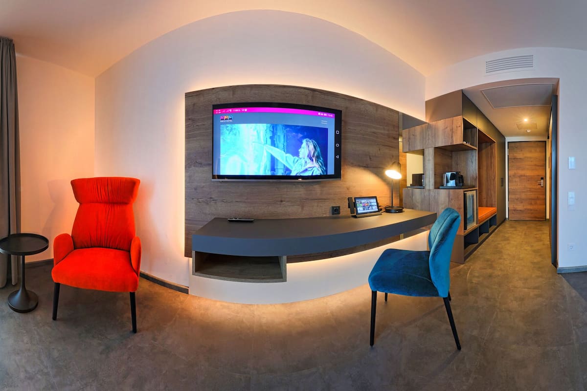 A modern hotel room with a bed, a desk, a TV, and a seating area.