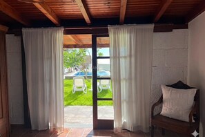 A room with a wooden ceiling, white walls, a door with white curtains, a wooden bench with a cushion, and a view of a swimming pool and chairs outside.