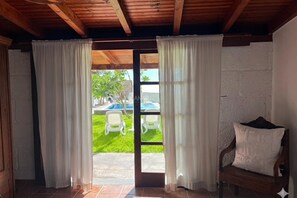 Een kamer met een houten plafond, witte muren, een deur met witte gordijnen, een houten bank met kussen en uitzicht op een zwembad en stoelen buiten.