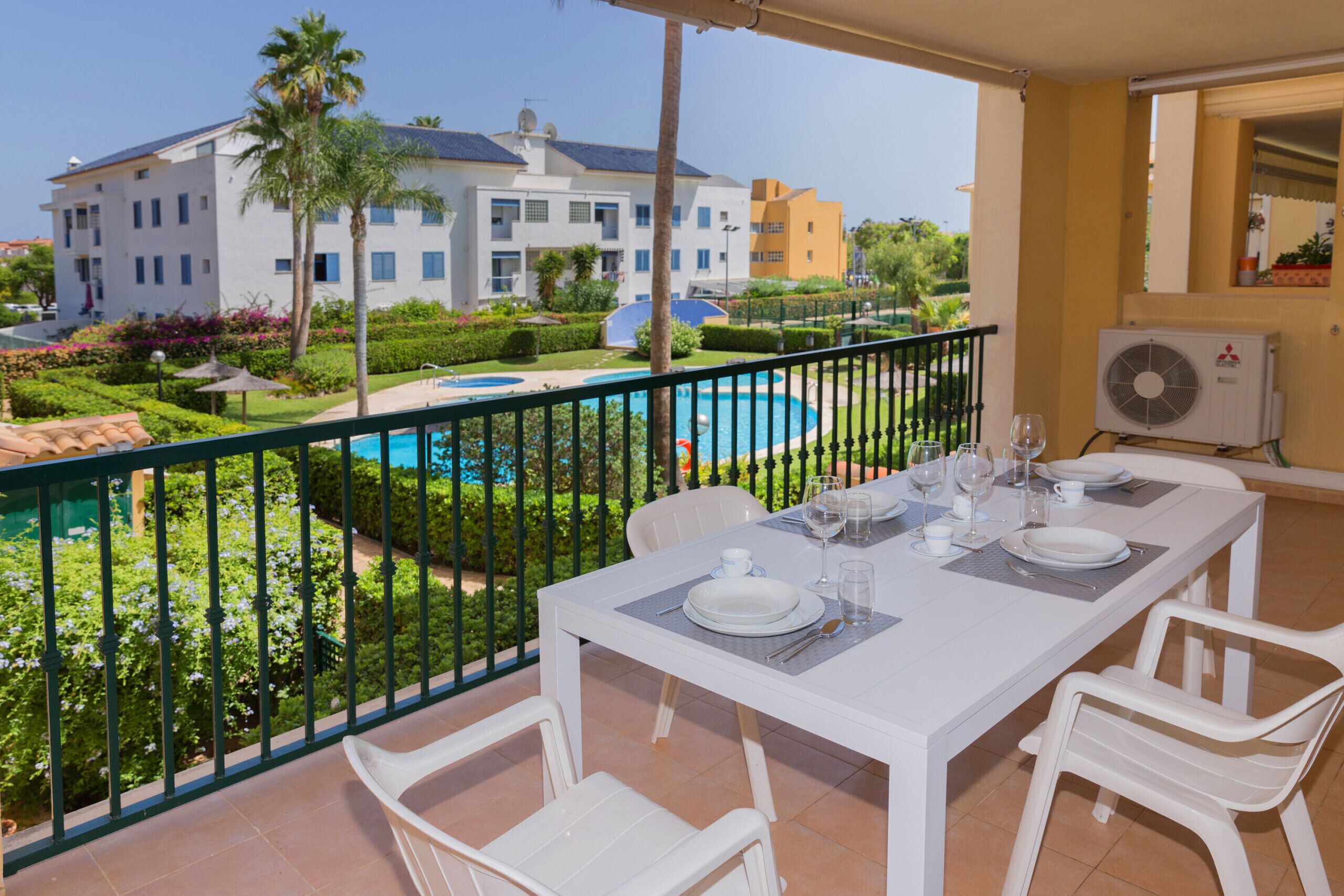 A balcony with a dining table set for four, overlooking a swimming pool and a landscaped garden.