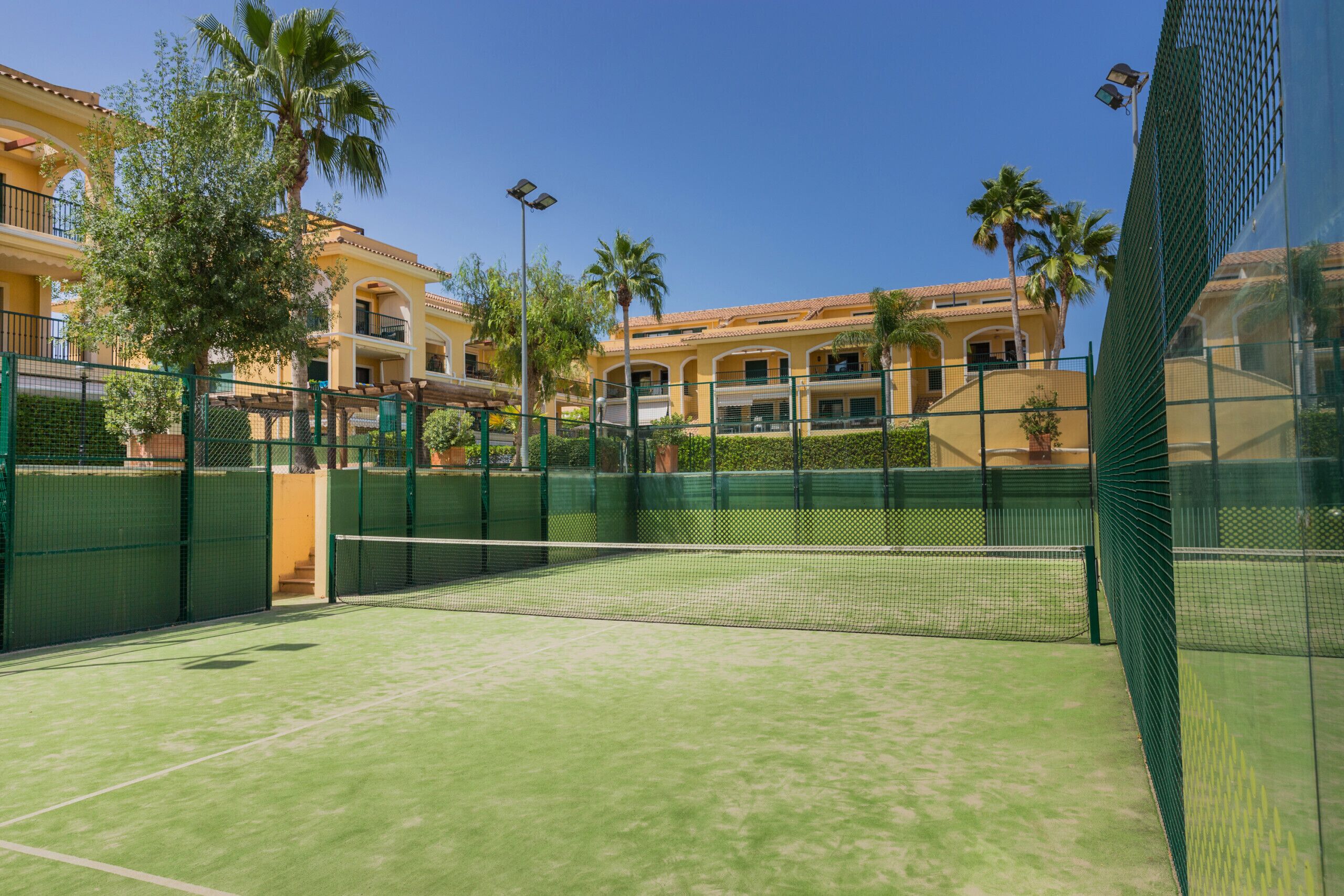 Una cancha de tenis con una red, rodeada por una valla verde, y edificios al fondo.