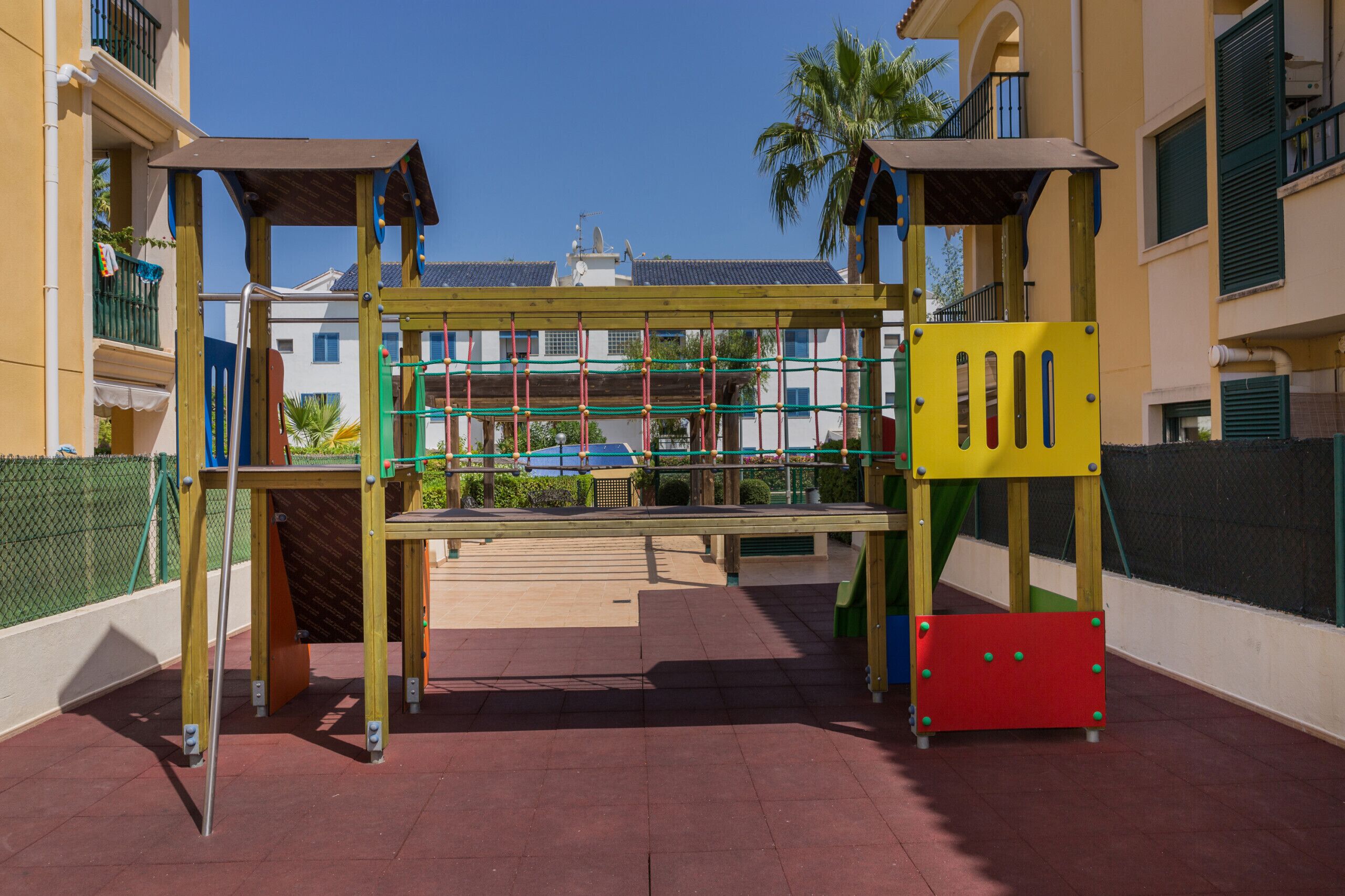 A playground with climbing structures and a slide, surrounded by buildings and a fenced area.