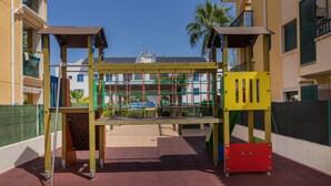 A playground with climbing structures and a slide, surrounded by buildings and a fenced area.