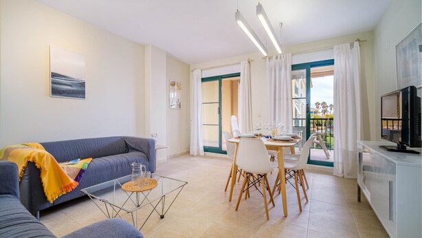 A modern living room with a sofa, dining table, and chairs. There is a kitchen area with a TV and cabinets.
