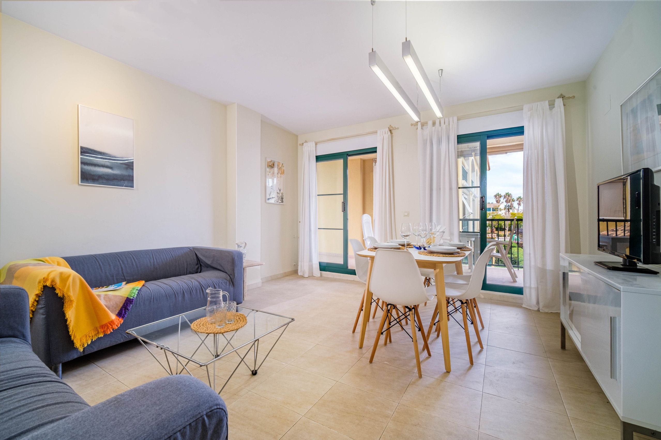 A modern living room with a sofa, dining table, and chairs. There is a kitchen area with a TV and cabinets.