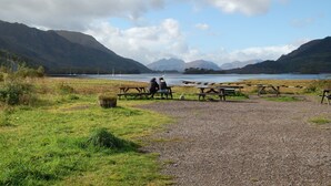 A lakeside picnic area with benches and picnic tables, surrounded by mountains and a clear sky.