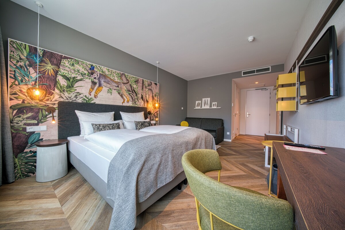 A modern hotel room with a large bed, a desk, a chair, a television, and a wall mural.