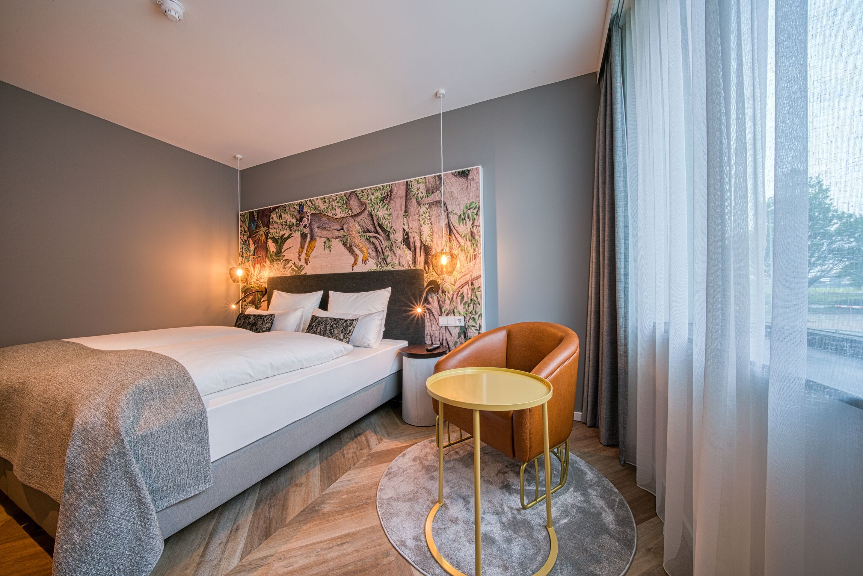 Une chambre d’hôtel moderne avec un grand lit, une petite table ronde et une vue sur l’extérieur à travers des rideaux légers.