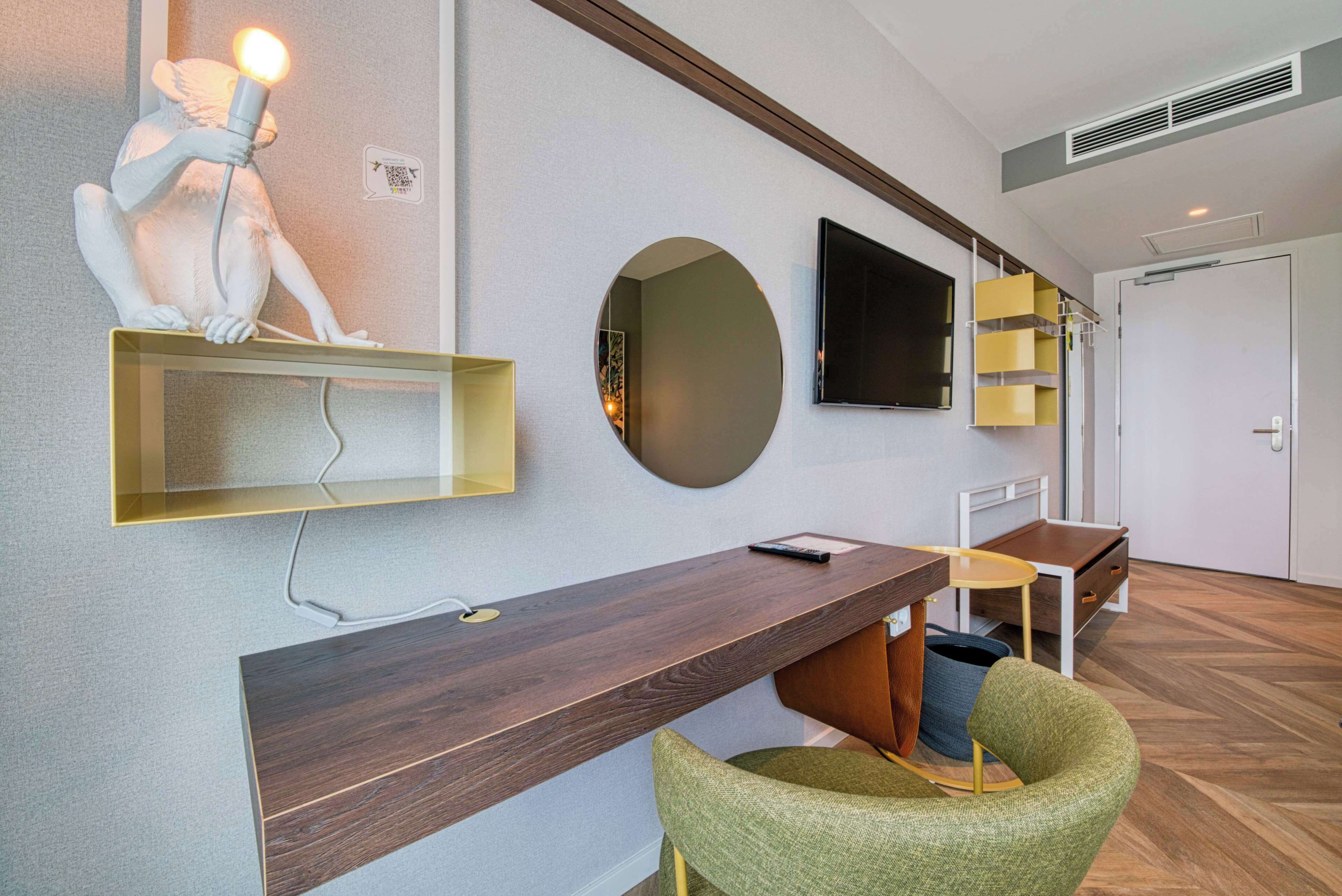Ein modernes Hotelzimmer mit einem Holzbüro, einem grünen Sessel, einem großen Spiegel und einem flachbildfernseher an der Wand.