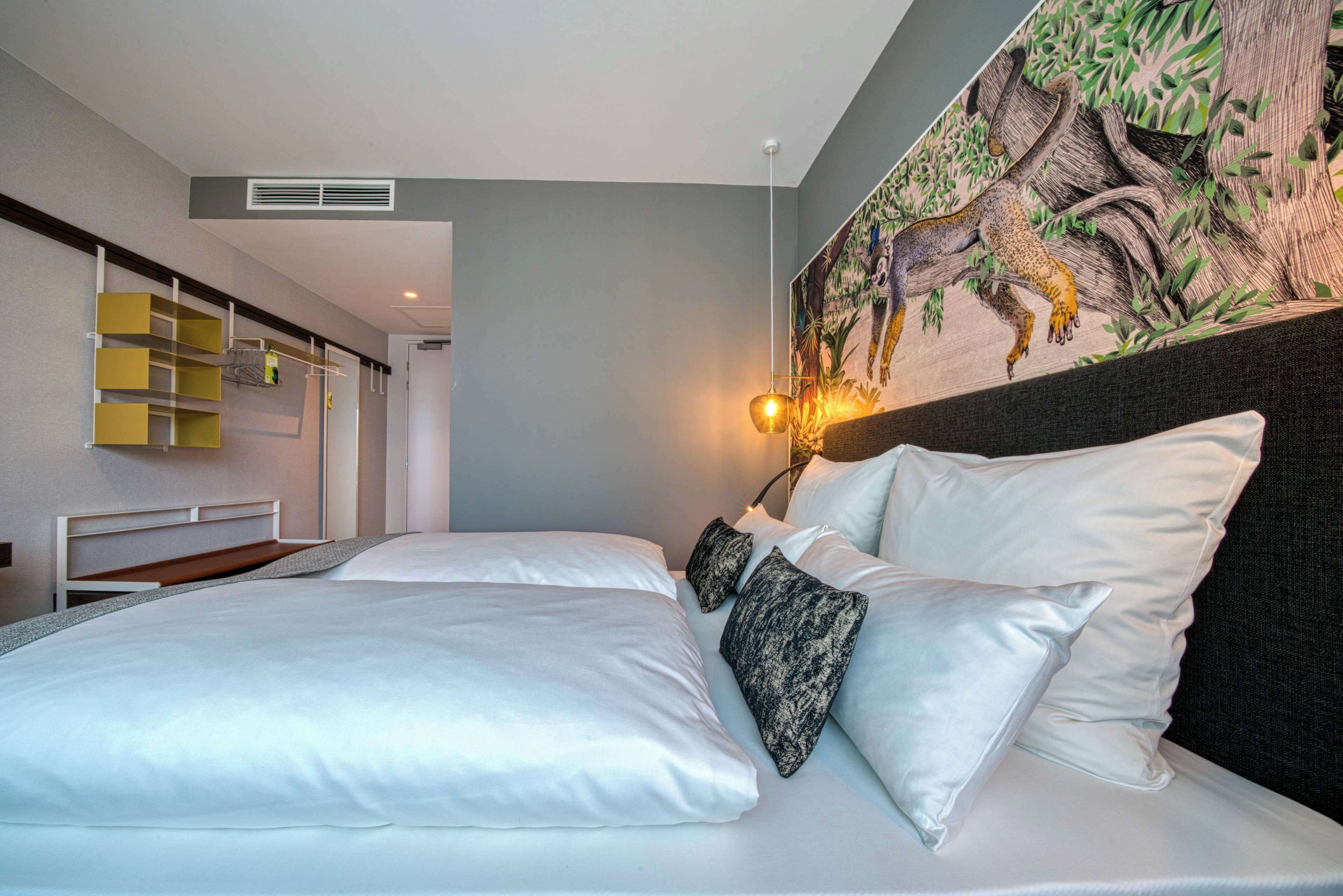 Une chambre d’hôtel avec un lit, du linge de lit blanc, une tête de lit ornée d’un tableau représentant un léopard et une étagère fixée au mur.