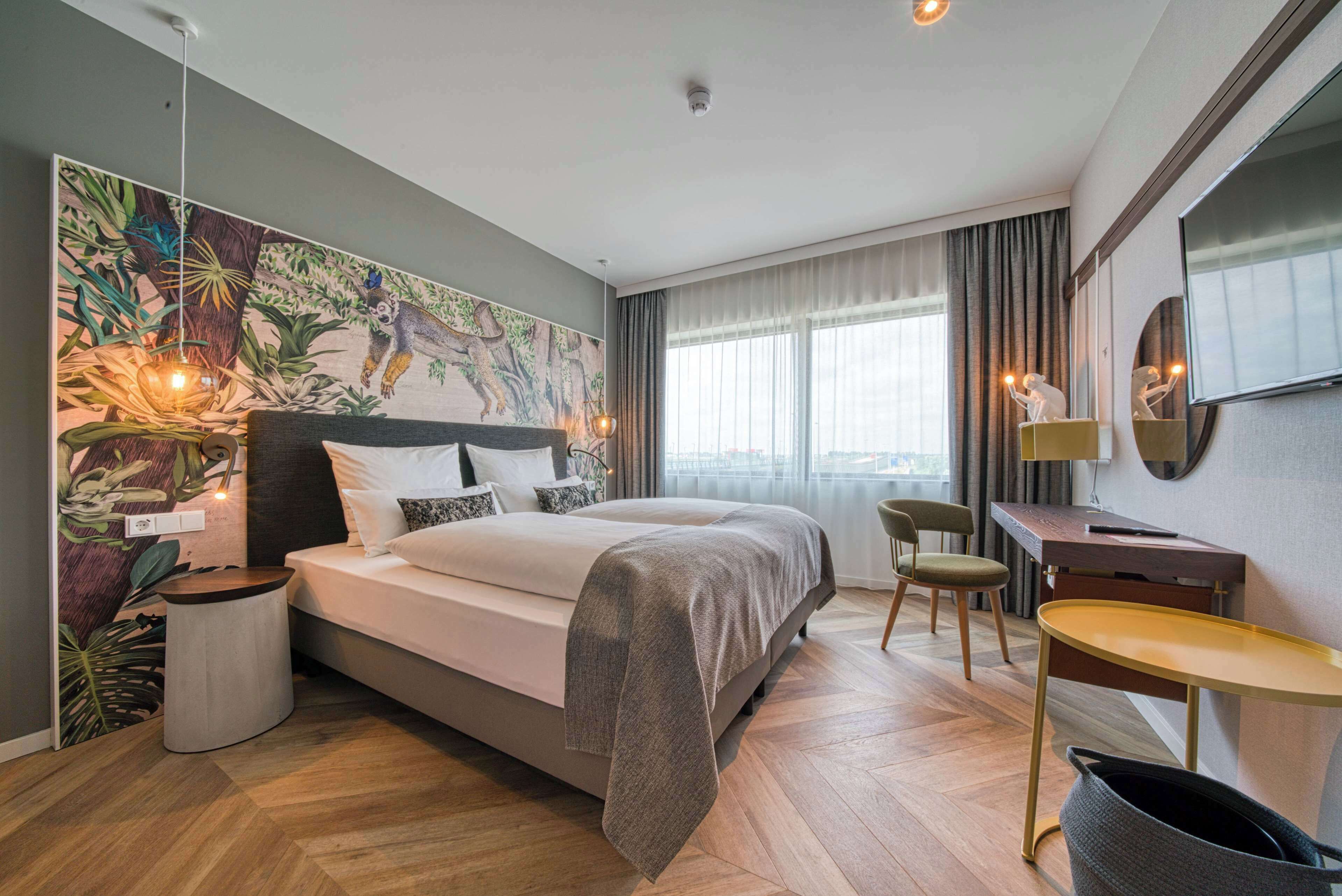 Una habitación de hotel moderna con una cama grande, un escritorio y una silla. Se observa un mural grande en la pared, una ventana con cortinas y una planta en la mesita de noche.